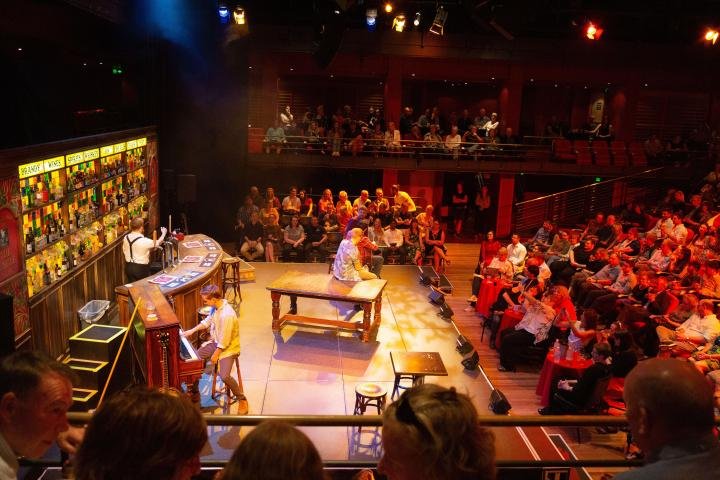 An indoor theater scene with a bar stage setup, featuring performers and an audience in a vibrant, lit environment. The stage is adorned with a bar counter and colorful bottles, while spectators are seated at tables and around the performance area.