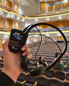A hand holding a wireless receiver and headphones in a concert hall, showing a display labeled 'RX-12' and 'City Recital Hall' in the background.